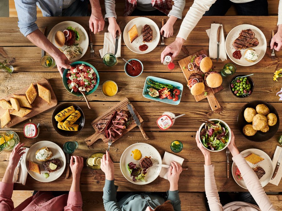 Sechs Menschen sitzen an einem Tisch und bedienen sich an dem Essen, was auf dem Tisch steht. Es gibt eine Auswahl an Brot, Salat und Fleisch.