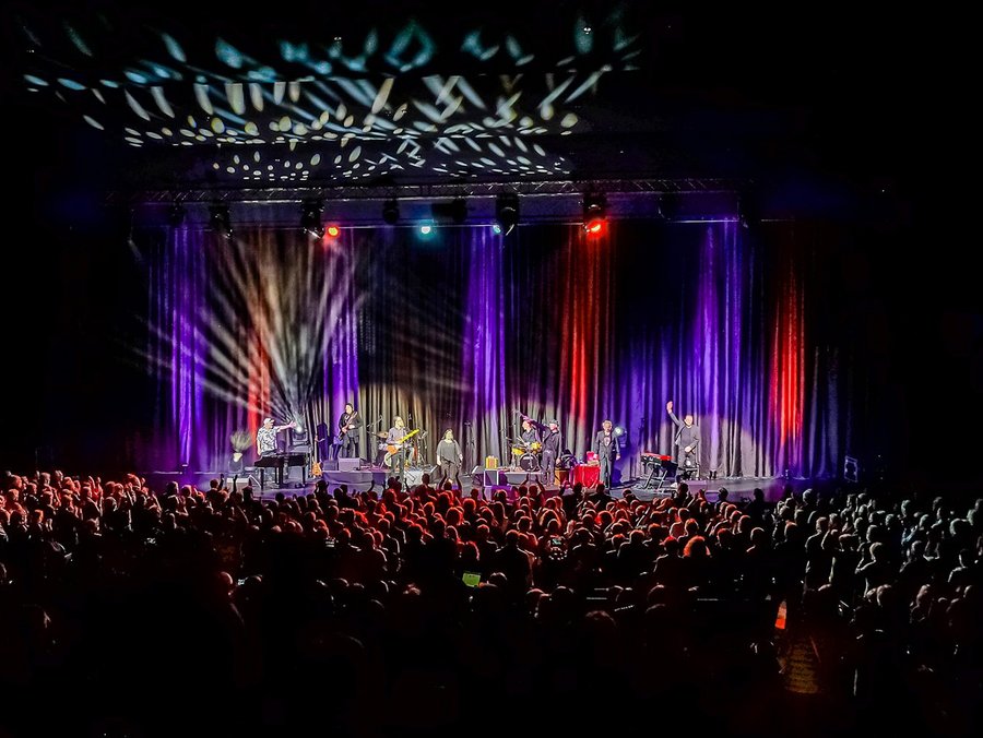 Auf einer Bühne spielt eine Band. Die Bühne ist rot und lila ausgeleuchtet. Im Vordergrund sind viele Menschen, die der Band zuhören.