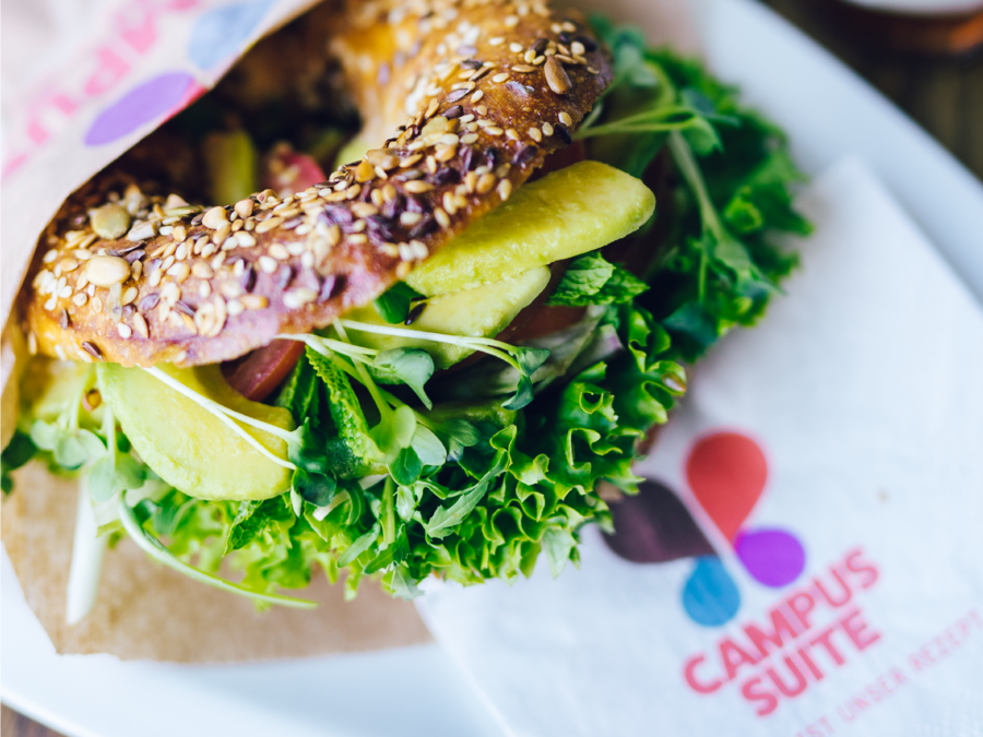 Der Bagel mit Körnerm ist belegt mit Avocado und Salat und liegt auf einer weißen Service mit dem Schriftzug der Campus Suite.