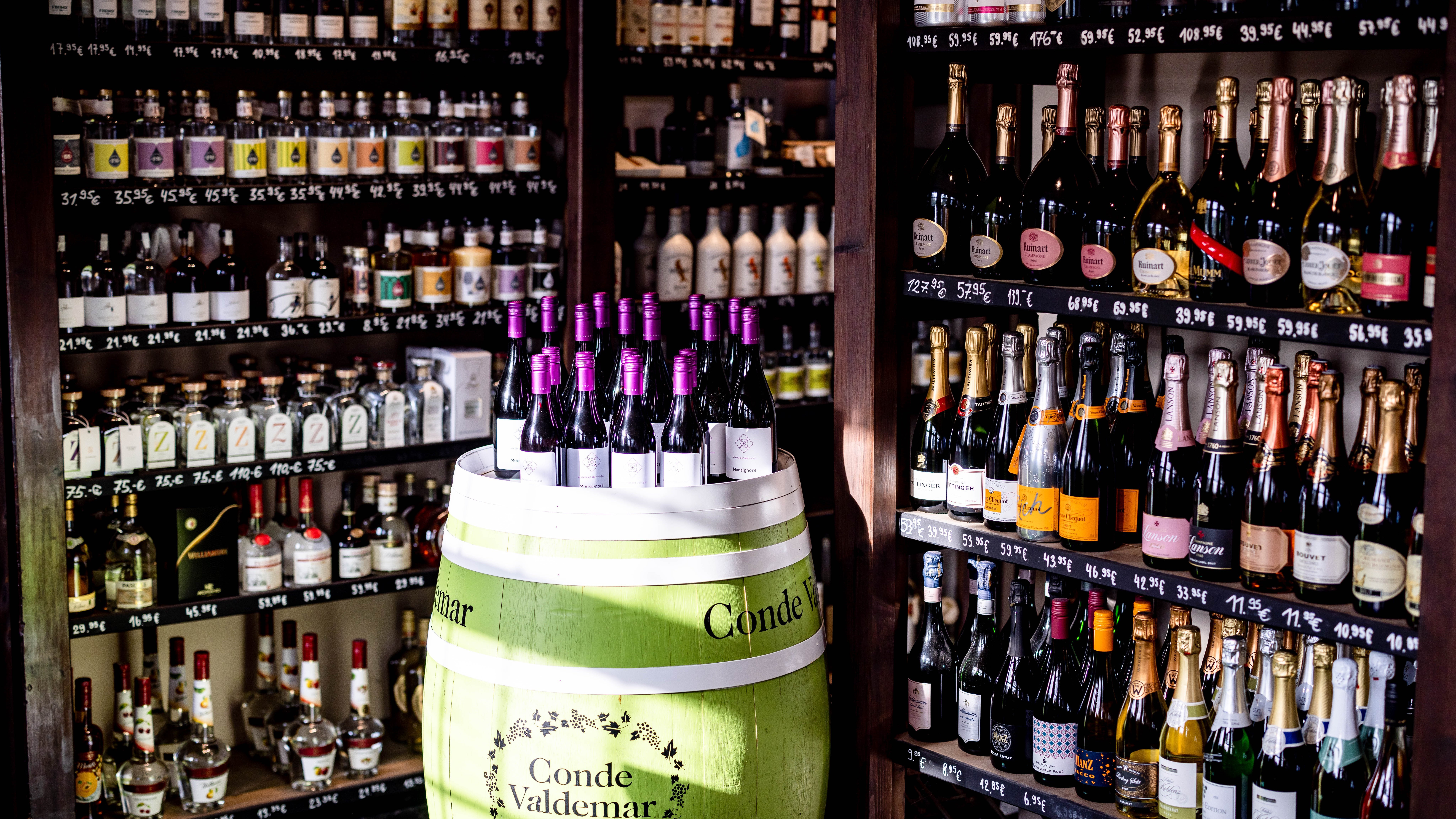 Ein über Eck gefülltes Weinregal Deckenhoch. Davor steht ein grün/weißes Weinholzfass mit vielen Flsschen Wein einer gleichen Sorte