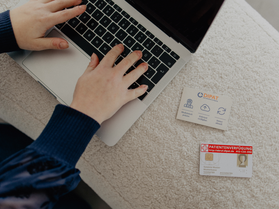 Ein Laptop der auf dem Bild nur zur Hälfte zu sehen ist. darauf sind 2 Hände die auf der Tastatur schreiben, rechts im Bild ist eine Dipat Visitenkarte und eine Patientenverfügungskarte in Form einer Scheckkarte