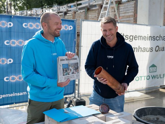 Zwei Männer auf einer Baustelle, hinter ihnen Bauplakate. Der Mann links im cyanfarbenen coop Pullover hält eine aktuelle Ausgabe der Kieler Nachrichten hoch. Der Mann rechts im dunkelblauen coop Pullover hält die röhrenförmige, kupferfarbene Zeitkapsel in den Händen.