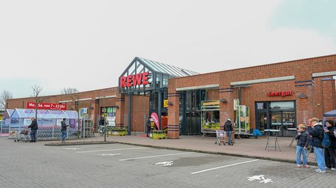 Die Außenfront des Supermarktes aus Klinker trägt das große REWE-Logo. Menschen sind im Vordergrund zu sehen.
