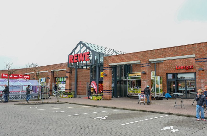 Die Außenfront des Supermarktes aus Klinker trägt das große REWE-Logo. Menschen sind im Vordergrund zu sehen.