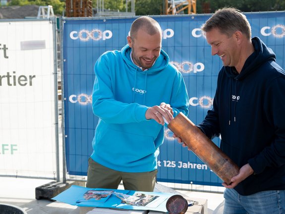 Zwei Männer auf einer Baustelle, hinter ihnen Bauplakate. Der Mann links im cyanfarbenen coop Pullover steckt etwas in eine kupferfarbene, röhrenförmige Zeitkapsel, die ein Mann rechts in einem dunkelblauen coop Pullover hält.