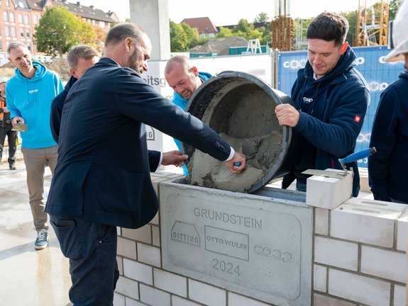 Auf der Baustelle stehen zwei Männer hinter einem Grundstein und halten einen großen Behälter mit Beton schräg über die obere Öffnung des Grundsteins. Ein Mann im Anzug, der vor dem Grundstein steht, nimmt mit einem Schaber Beton aus dem Behälter und füllt damit damit den Grundstein. Links im Hintergrund sieht man eine Person im cyanfarbenen coop Pullover, die sich neugierig guckend vorbeugt.