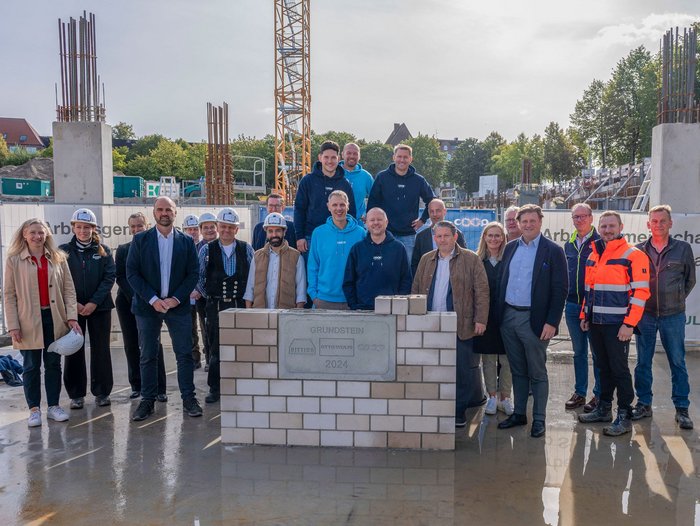 Neben und hinter einem grauen Grundstein stehen auf einer Baustelle im Gegenlicht viele Menschen und schauen in die Kamera. Ein Gruppenfoto zur Grundsteinlegung.