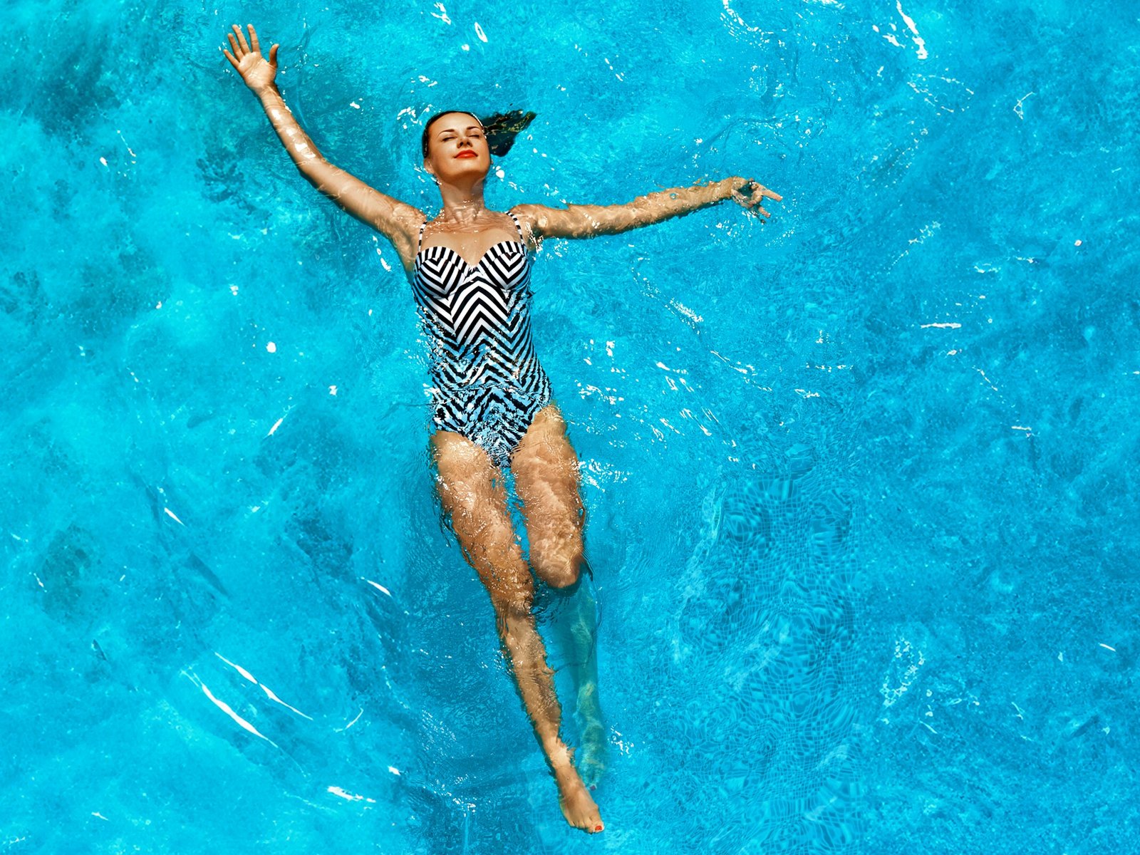 Eine Frau im gemusterten Badeanzug schwimmt auf dem Rücken in blauem Wasser. 