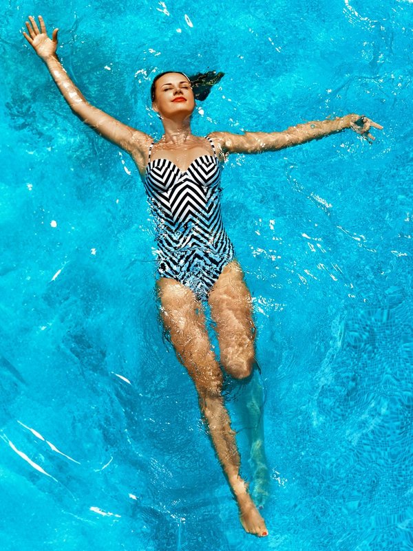 Eine Frau im gemusterten Badeanzug schwimmt auf dem Rücken in blauem Wasser. 