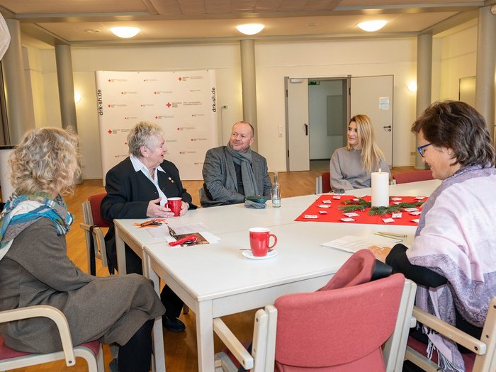 Fünf Personen sitzen an einem großen, weißen Tisch. Im Hintergrund steht Fotowand des DRK.