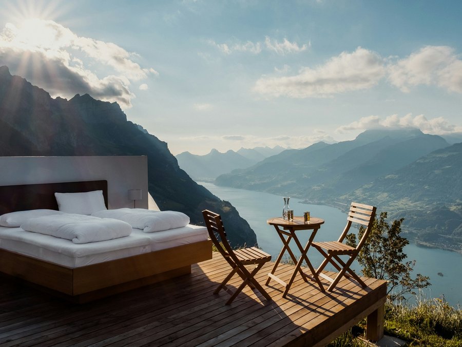 Ein Doppeltbett mit weißer Bettwäsche steht auf einer Terrasse aus Holz mit Aussicht auf Berge und einen See. 