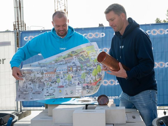 Zwei Männer falten ein großes Wimmelbild zusammen. Man sieht darauf die coop Geschichte. Der Mann links steht hinter dem WImmelbild in einem cyanfarbenen Pullover, der Mann rechts ist komplett zu sehen und trägt einen dunkelblauen coop Pullover.