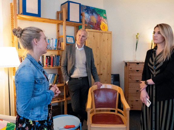 Zwei Frauen und ein Mann stehen in einem gemütlichen Raum mit Bücherregalen und Schränken im Hintergrund. Die Frau links spricht mit einer Geste der Hände, während der Mann in der Mitte mit verschränkten Armen aufmerksam zuhört. Die Frau rechts blickt interessiert in Richtung der Sprecherin, hält dabei ein Handy locker in den Händen.