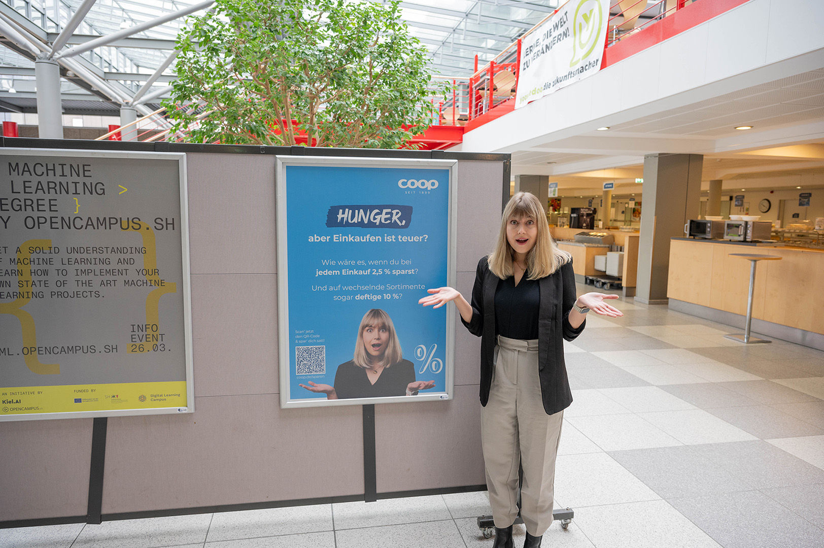 Paulina, Mitarbeiterin der coop, steht neben einem blauen Werbeplakat in der Mensa der CAU Kiel.