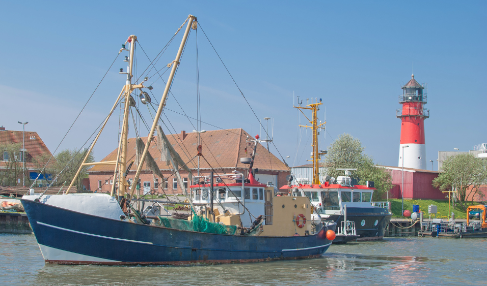 Ein Fischkutter fährt im Wasser des Büsumer Hafens. Im Hintergrund steht der Büsumer Leuchtturm vor blauem Himmel.