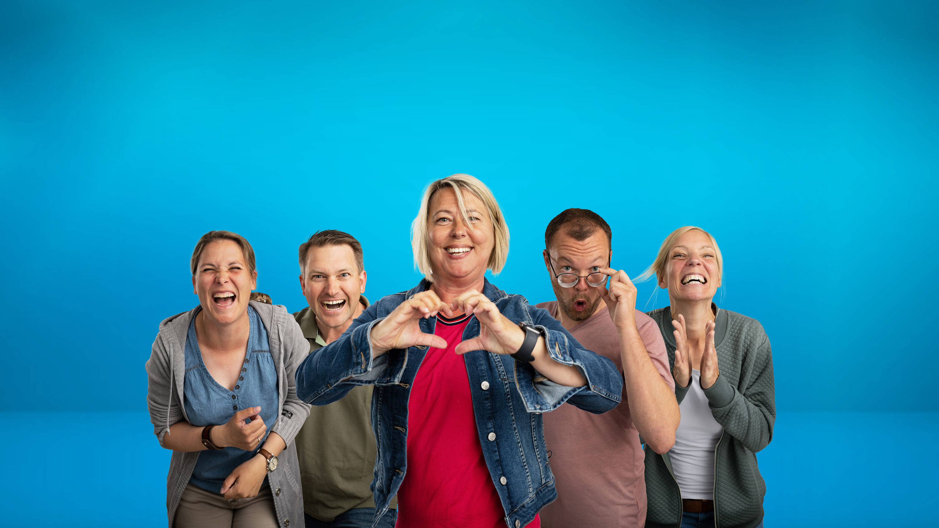 Eine fröhliche Gruppe von fünf Personen vor einem leuchtend blauen Hintergrund. In der Mitte steht eine Frau mit blonden Haaren, einer Jeansjacke und einem roten Oberteil, die ein Herz mit ihren Händen formt. Links von ihr stehen eine Frau mit kurzen braunen Haaren, die herzlich lacht, und ein Mann mit dunklem Haar, der breit grinst. Rechts von ihr steht ein Mann mit Bart, der seine Brille leicht herunterzieht und in die Kamera schaut, sowie eine Frau mit kurzen, hellblonden Haaren, die freudig in die Hände klatscht.