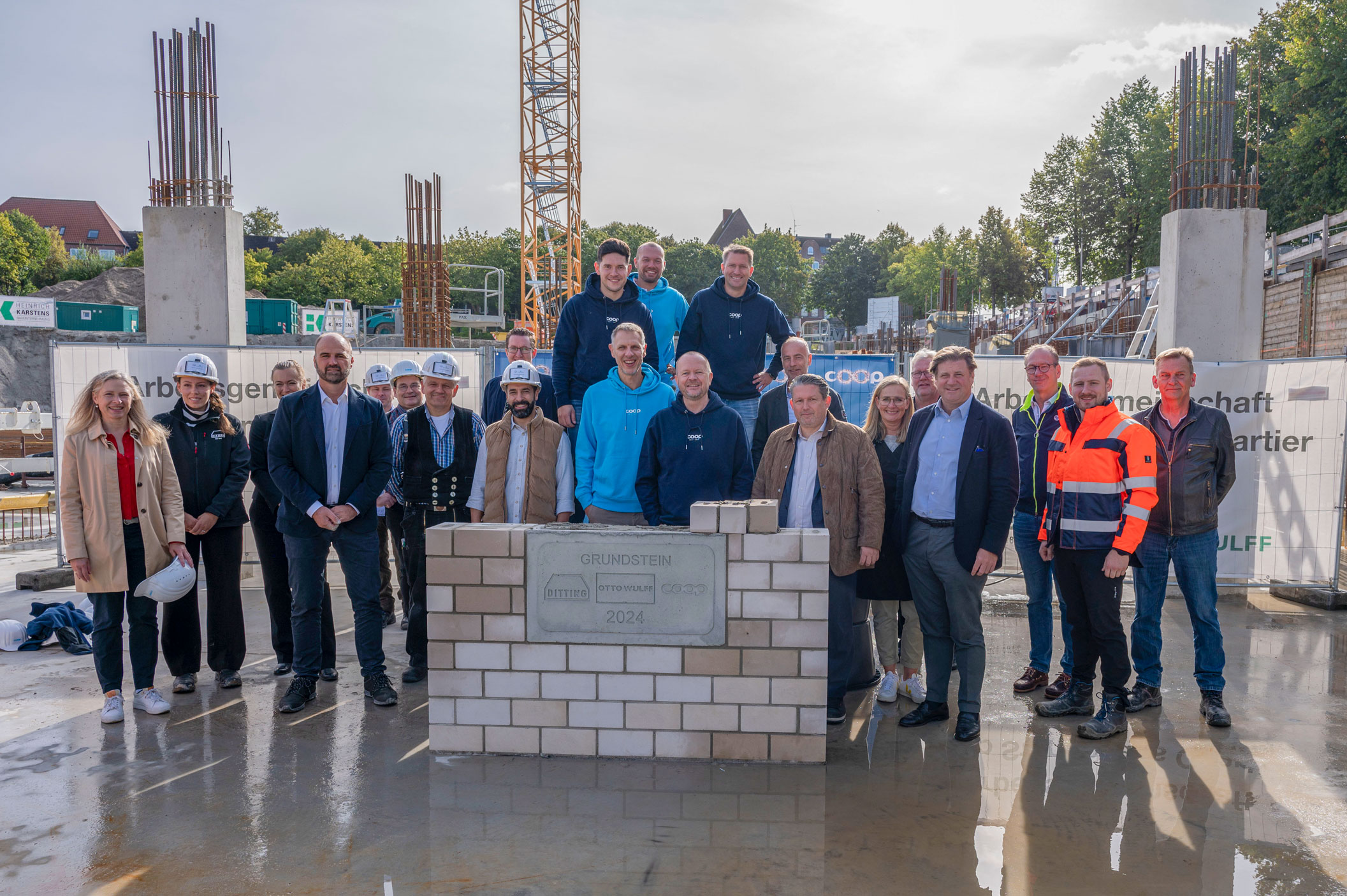 Neben und hinter einem grauen Grundstein stehen auf einer Baustelle im Gegenlicht viele Menschen und schauen in die Kamera. Ein Gruppenfoto zur Grundsteinlegung.