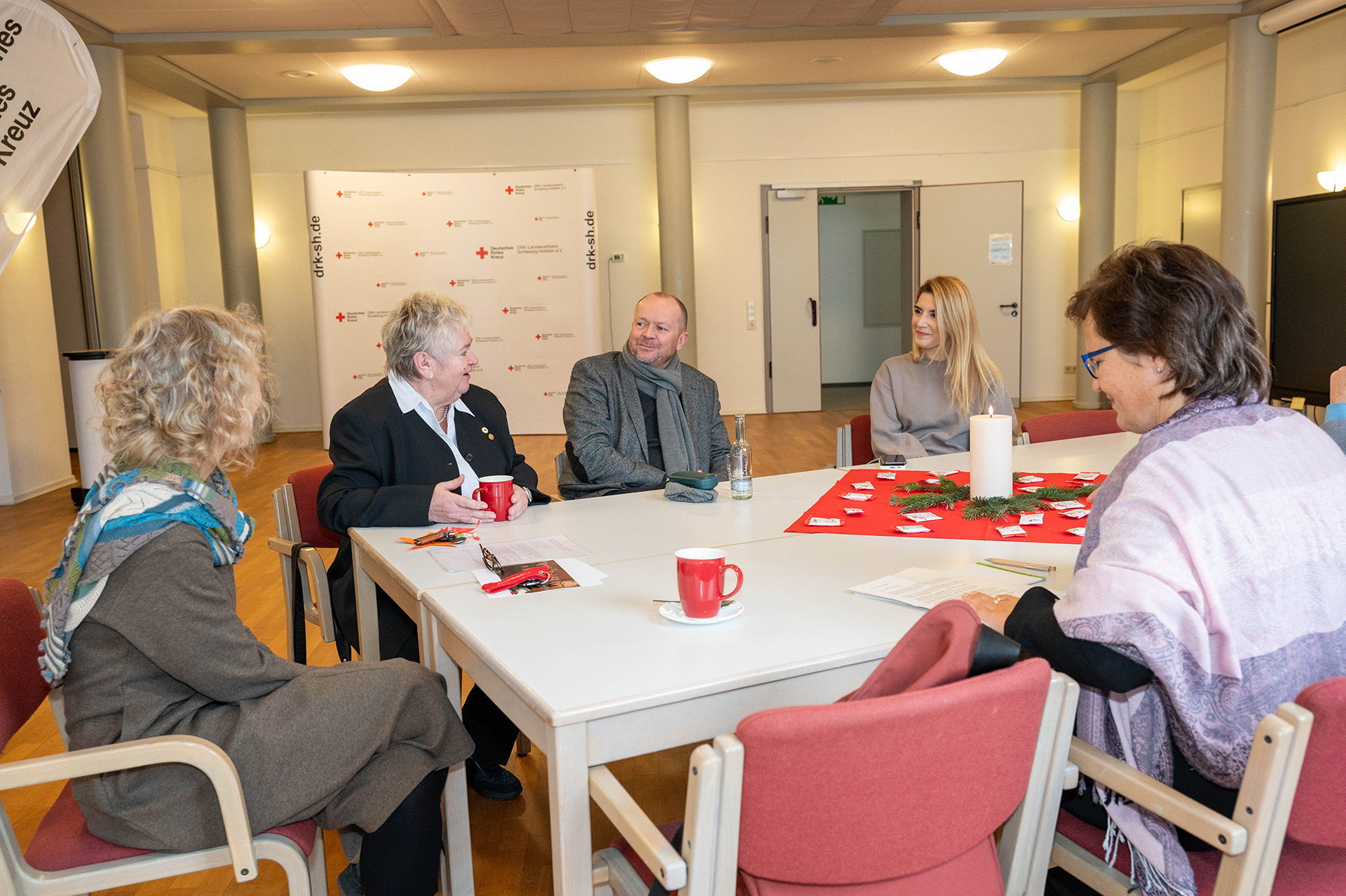 Fünf Personen sitzen an einem großen, weißen Tisch. Im Hintergrund steht Fotowand des DRK.